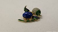 Brooch showing blue enamel wings, lapis stone body, green enamel head and tail, and rhinestone-studded head detail lying on white fabric