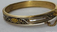 Close-up image showing gold-tone bangle bracelet with black and gold ornamental designs and rectangular mother of pearl inlay. Safety chain and box clasp visible.