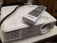 Side-front view of white BenQ projector with power cable and remote control on top.