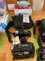 Three camcorders and a Roots camera bag arranged on a wooden table; Sony camcorder with 67x zoom and Panasonic camcorder behind it; Canon FS200 camcorder on box with power adapter and Roots bag in front