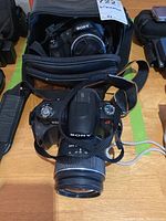 Sony Alpha DSLR A230 camera front with lens and strap, placed in front of a camera bag containing another Sony camera.