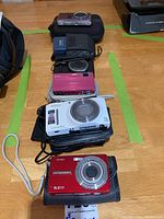 Five cameras laid on table in soft cases, front to back: red Kodak, white Nikon, pink Sony, black Canon, purple Samsung
