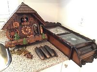 Overview of two clocks on countertop: chalet cuckoo clock on left, rectangular wall chime clock on right, with weights and pendulum components visible