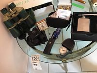 Overview photo of lot items on glass table including binoculars, mini binoculars in case, pocket watch with cutter, flask and lighter set, folding knife, small clock, and a small black binoculars