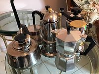 Photo showing five metal coffee makers and kettles on a glass table with reflective surface. Various shapes including hexagonal and rounded models with black plastic handles and gold knobs on top of some lids.