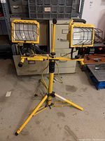 Front view of dual yellow Home Light construction lights with metal wire guards attached to adjustable yellow tripod stand on concrete floor.
