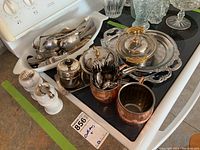 Photo showing metal trays with ornate handles, glass containers with lids, copper cups, stainless steel cutlery, and salt and pepper shakers arranged on a counter.