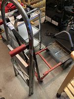 Photo of a metal dolly with red handles and wheels in a garage setting, showing some rust on the metal surfaces.