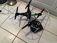 Full view of black Alile quadcopter drone with protective propeller guards and corresponding remote controller on tiled floor.
