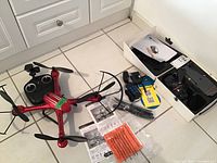 Red Propel X15 quadcopter on floor next to its remote control, extra blades, propeller guards, USB cable and instruction sheets