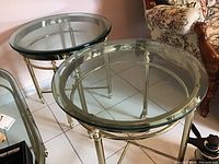 Two matching round end tables with thick glass tops and gold metal frames, placed on tiled floor near floral upholstered furniture.