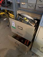 Metal two drawer filing cabinet opened showing tools inside including a circular saw, saw blades and hand tools.