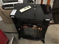 Full view of black decorative electric fireplace with glass front and brass handle showing interior faux logs and fire effect, along with remote placed on top.