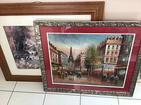 Two framed prints leaning against a wall: one with a Paris street scene including the Eiffel Tower, the other with a nature landscape showing a stream and trees.