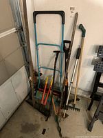 Photo shows a collection of gardening tools leaning against the wall including a vintage manual push lawn mower, rakes, clippers, and weed pullers.