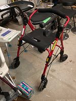Front and side view of the red rollator walker with seat and backrest, showing four wheels and foam-padded handles with brakes.