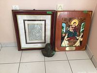 Framed silver embossed religious artwork, wooden framed tile art, and pope bust statue placed side by side on floor.