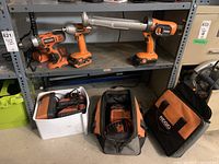 Wide view of the shelf showing Ridgid cordless impact driver, cordless drill, cordless caulk/adhesive gun, battery chargers, and two Ridgid tool bags on the floor