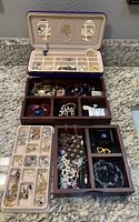 Open jewelry case on countertop showing top compartment with assorted earrings, rings, lapel pins and pearl bracelets