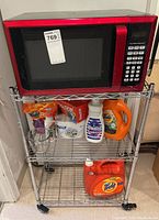 Red Hamilton Beach microwave sitting on top shelf of a chrome metal wire rack with laundry detergents and cleaning supplies visible on two lower shelves.