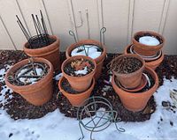 Photo showing clay pots of various sizes partially filled with soil, dried plant material, and some snow. Several pots are stacked or nested. Two metal plant stands are visible.