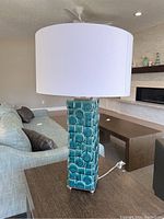 Side view of the rectangular blue ceramic lamp base with raised circular and square patterns, topped with a wide white lampshade, placed on a dark wood side table in a living room.