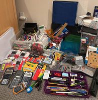 Wide view showing an assortment of office supplies including multiple calculators, Sharpie marker packs, Crayola markers, various pens and pencils, glue sticks, and desk tools scattered on the floor and in containers.
