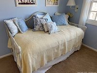 Daybed with white metal vintage frame, light yellow bedspread, and blue and floral pillows in a carpeted room with blue walls.