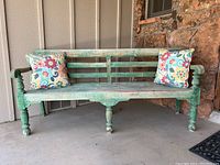 Front view of a vintage wooden bench painted in distressed green with two floral patterned pillows on a porch.