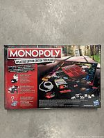 Back of unopened Monopoly Cheaters Edition box showing detailed gameplay elements and components including cheat cards and handcuffs.