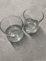 Two thick bottom scotch glasses placed on a grey surface, showing clear views of their shape, thickness of base, and transparency of glass.
