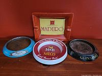 Top view of four vintage advertising ashtrays placed side by side on red surface, showing front advertising logos with good condition.