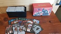 Photo showing a black card storage box filled with Magic The Gathering token cards and a red Magic The Gathering box used for card storage. Several scattered token cards showcase a variety of artwork and colors.