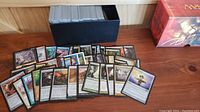 Photo showing a collection of Magic The Gathering cards spread out in front of a black storage box on a wooden surface next to a red Magic The Gathering game box.