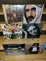 Photo showing six vinyl record albums standing/sitting on a wooden surface. Visible artists include Frank Zappa, Adele (21), Ben John (Babylon), Michael Jackson (Off The Wall), The Moody Blues, and an additional album 'Say Down Stranger'.