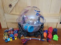Image shows clear plastic battle dome with black base, multiple Beyblade tops, and parts including launchers and disks arranged in front of dome on wood floor