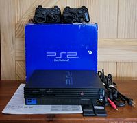 Front view of PlayStation 2 console with original box behind, two controllers on top, cables and memory cards in front, instruction manual.