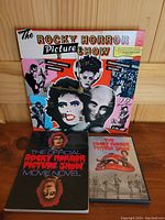 Photo of three Rocky Horror Picture Show items: a wall clock with character artwork, a DVD movie, and a 1980 movie novel book.