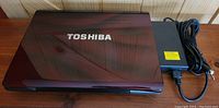 Closed top lid of the Toshiba laptop with power adapter and cord beside it on wooden surface, showing dark maroon color and Toshiba logo.