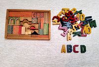 Wooden building blocks with printed architectural patterns arranged inside a wooden box with missing top, with colorful alphabet letters next to it, some letters appear broken or missing.