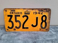 Front view of 1942 Ontario metal car license plate showing yellow background, black numbers '352 J8', black text 'ONTARIO', crown symbol, and year '1942'. Some rust and paint wear visible around edges and mounting holes.