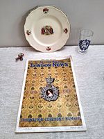 Photo showing a white commemorative plate with coronation motifs, a vintage glass, a lapel pin with ribbon, and an Illustrated London News magazine with golden decorative cover.