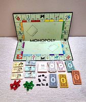 1961 Monopoly game board folded open with metal playing pieces, houses, hotels, paper money and cards displayed in front