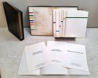 Photo of 8 white booklets with colored stripes on the covers and some loose pages, displayed with a black slipcase.