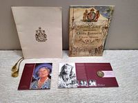 Shows all items included in the lot visible on a gray surface: cream hardcover book with gold tassel, a softcover book with royal crest, two booklet style folders with photographic portraits and coin attached