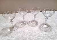 Four antique champagne glasses made of clear crystal displayed in a row on a cream textured surface, showing the etched floral and leaf pattern and thin stems.