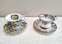 Two Royal Albert teacup sets side by side, one with multicolored roses on dark blue background and the other white with gray floral pattern.