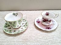 Two vintage Royal Albert teacup sets side by side on a white textured surface. One set has a green floral pattern, the other a pink rose floral pattern.