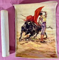 Photo of original oil painting on canvas showing matador with red cape facing charging black bull. The canvas is unframed and rolled beside wooden dowel.