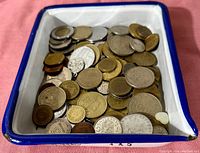 Coins of assorted types, sizes, and metals collected together inside a white enamel tray with a blue rim. Coins appear well circulated.
